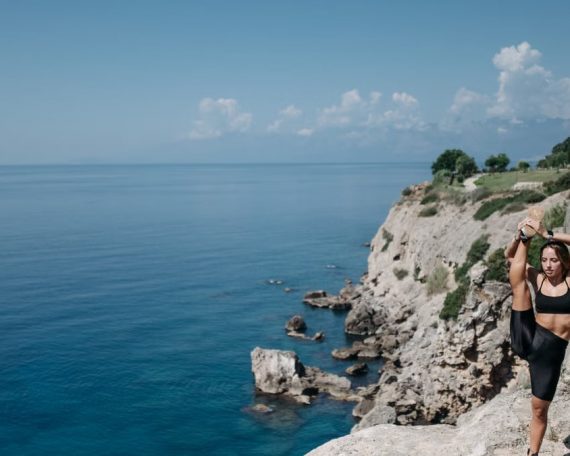 Woman in workout attire stretching on a cliff by the sea under a clear blue sky.